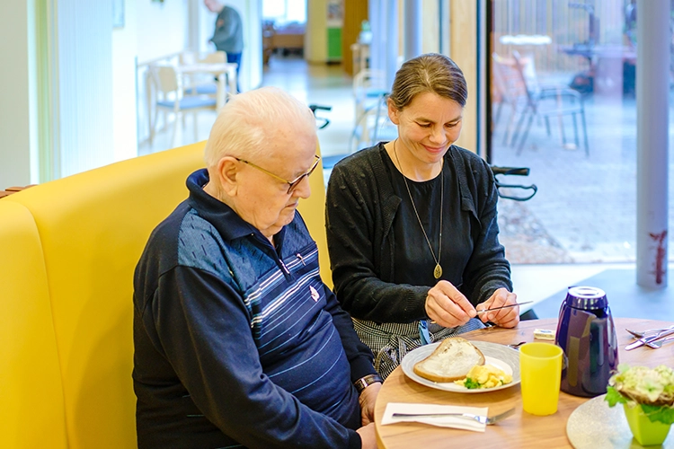Betreuung hilft älterem Mann beim Essen am Tisch