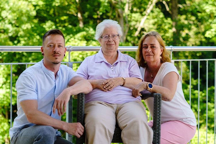 Seniorin sitzt mit zwei Angehörigen gemeinsam draußen auf einer Terrasse