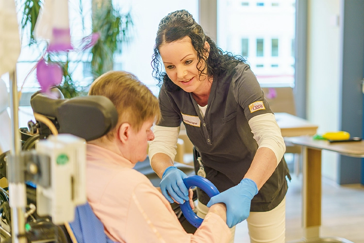 Therapeutin unterstützt Patientin im Rollstuhl bei einer Übung mit einem blauen Trainingsgerät.