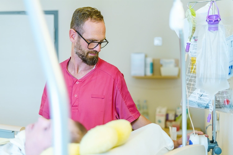 Pfleger in roter Kleidung kontrolliert medizinische Geräte neben einem Patientenbett.