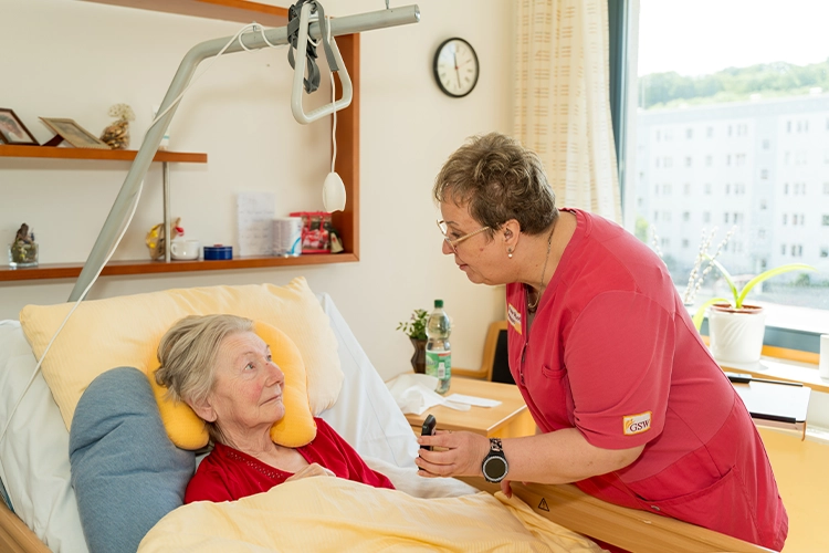Pflegekraft kümmert sich am Bett um eine ältere Patientin und spricht mit ihr