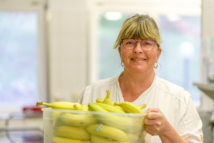 Kuechenmitarbeiterin hält Kiste mit Bananen und lächelt
