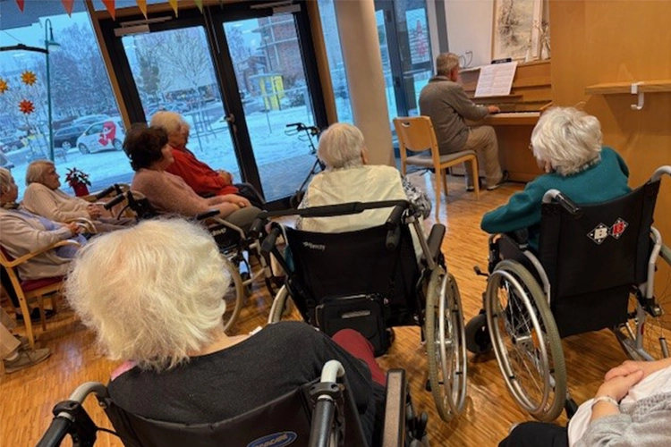 Ältere Menschen, teils in Rollstühlen, hören einer Person beim Klavierspielen zu.