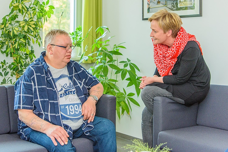Bewohner und Betreuerin sitzen auf einem Sofa im Gespräch umgeben von Zimmerpflanzen
