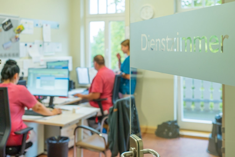 Blick durch Glastür mit Aufschrift „Dienstzimmer" auf zwei Pflegekräfte an Computerarbeitsplätzen