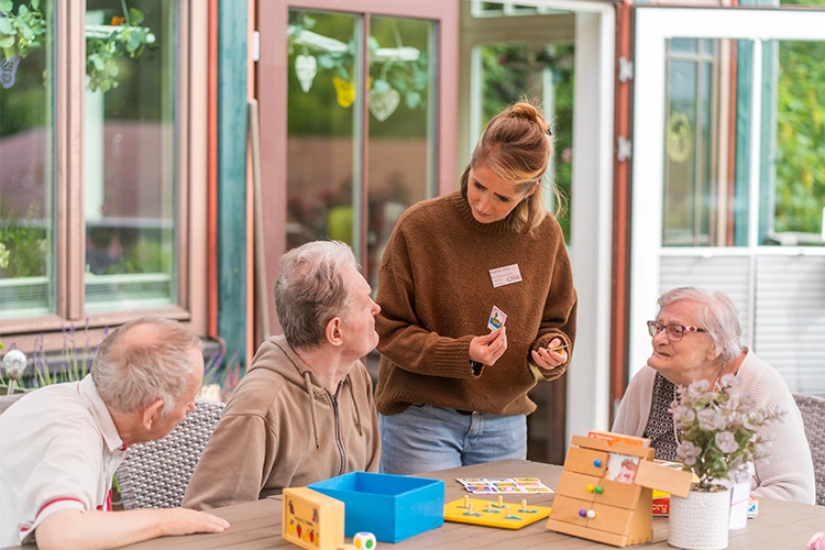 Betreuerin zeigt drei älteren Bewohnern Spielkarten am Tisch.