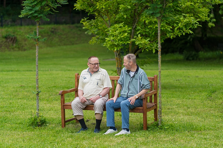 Zwei ältere Männer sitzen auf einer Holzbank im Park und unterhalten sich.