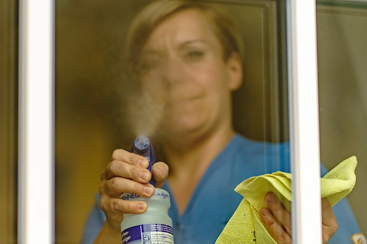 Frau in blauer Arbeitskleidung putzt eine Fensterscheibe mit Sprühflasche und Tuch