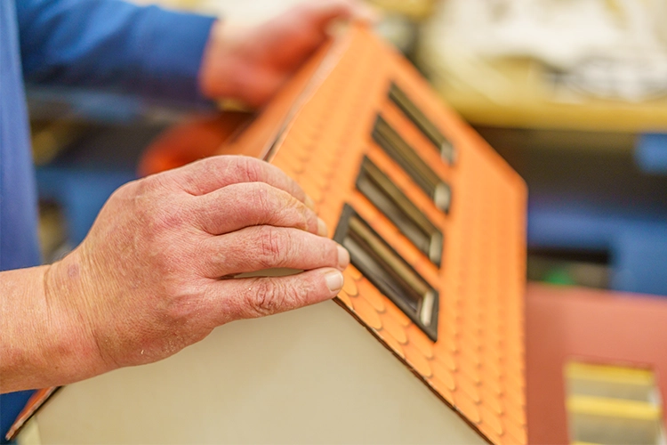 Nahaufnahme einer Hand, die ein orangefarbenes Holzhaus-Modell mit Nieten hält