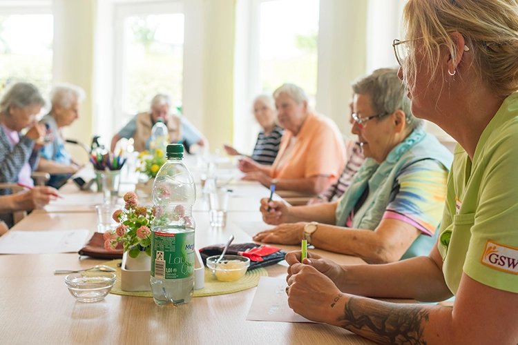 Betreuerin leitet eine Gruppe von Seniorinnen bei einer gemeinsamen Aktivität am langen Tisch