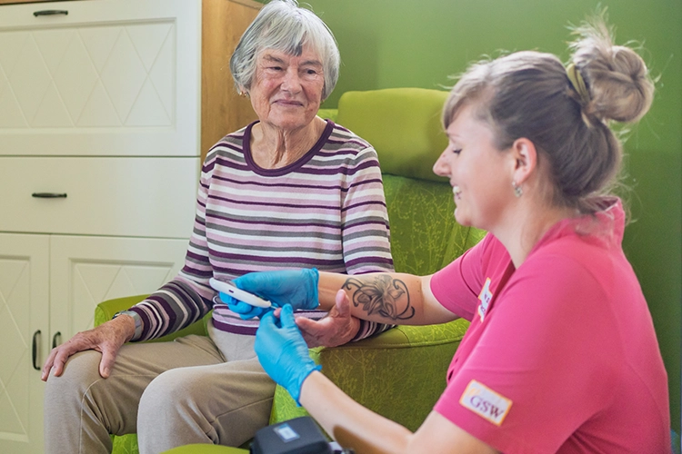 Pflegerin führt bei einer Seniorin im Sessel eine Blutzuckermessung durch