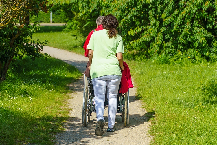 Pflegekraft schiebt eine Seniorin im Rollstuhl einen grünen Gartenweg entlang