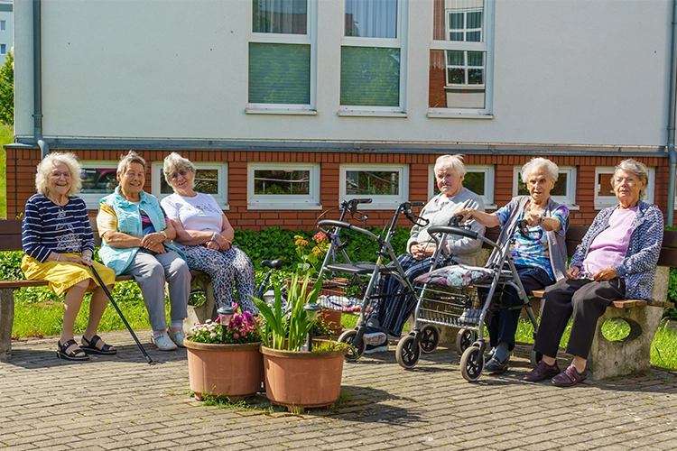 Sechs Seniorinnen sitzen gemeinsam auf Bänken im Freien vor dem Gebäude