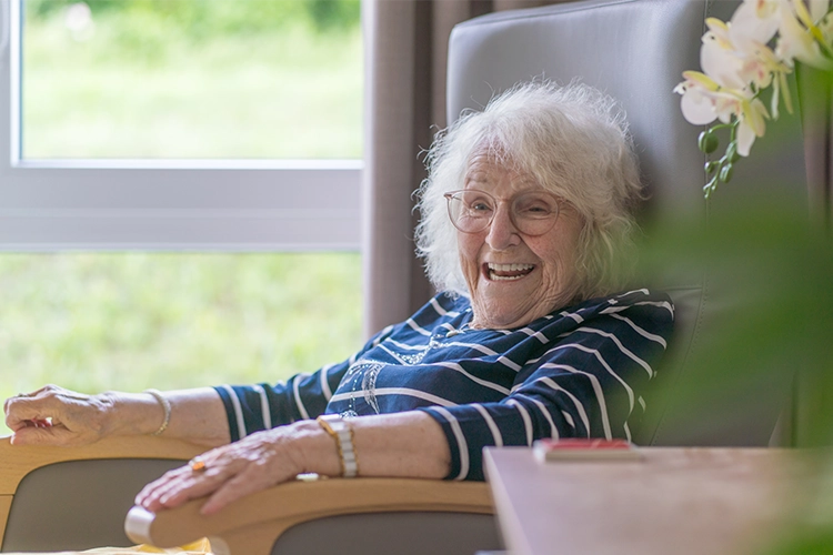 Ältere Dame sitzt lachend und entspannt in einem Sessel am Fenster