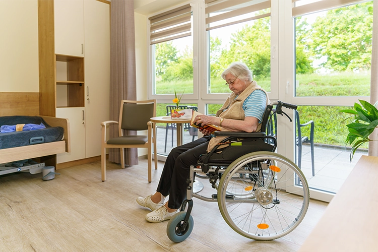 Seniorin im Rollstuhl liest ein Buch in einem hellen Zimmer mit Terrassenzugang