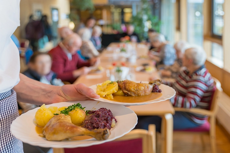 Servicekraft serviert zwei Teller mit Mittagessen an einem langen Tisch mit Senioren