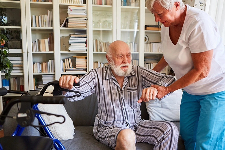 Ältere Frau hilft einem älteren Mann mit Bart beim Aufstehen vom Sofa neben einem Rollator