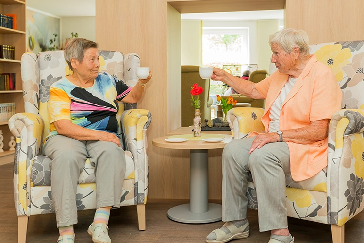 Zwei Seniorinnen sitzen in gemusterten Sesseln und stoßen mit Kaffeetassen an