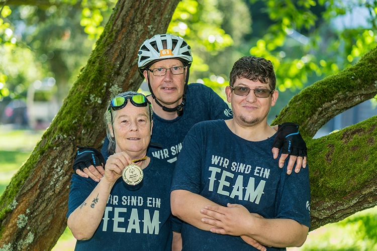 Drei Personen in blauen Team-Shirts stehen lächelnd im Park, eine hält eine Medaille