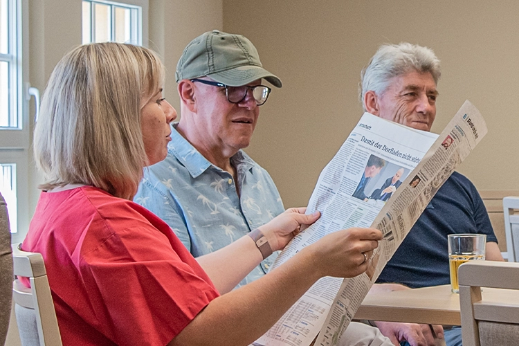 Betreuerin liest zwei älteren Männern laut aus einer Zeitung vor