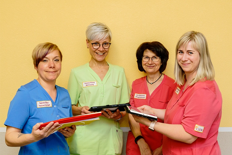 Vier Mitarbeiterinnen in Arbeitskleidung mit Namensschildern halten Unterlagen und Tablets