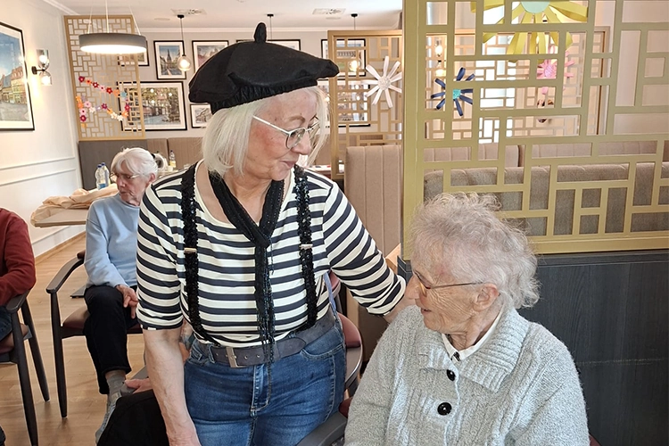 Frau mit schwarzer Baskenmütze und gestreiftem Oberteil unterhält sich mit einer sitzenden älteren Dame.