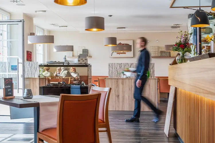 Gemütliches, helles Restaurant-Interieur mit Holzmöbeln, Pendelleuchten und einem Kellner in Bewegung.