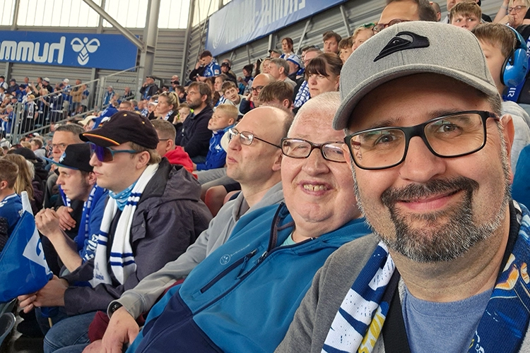 Selfie mehrerer Männer mit Fan-Schals auf einer Tribüne im Stadion.