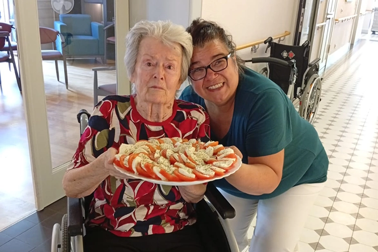 Ältere Frau im Rollstuhl und Betreuerin halten lächelnd eine Platte mit Tomate-Mozzarella.