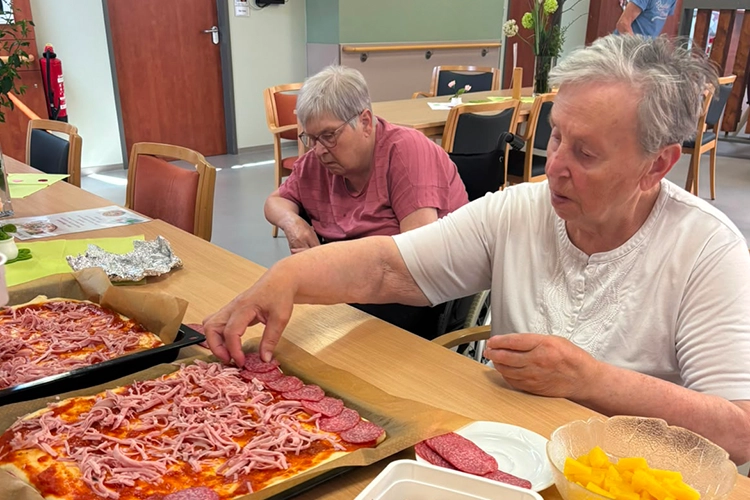 Zwei ältere Frauen belegen am Tisch ein Blech mit Pizza.