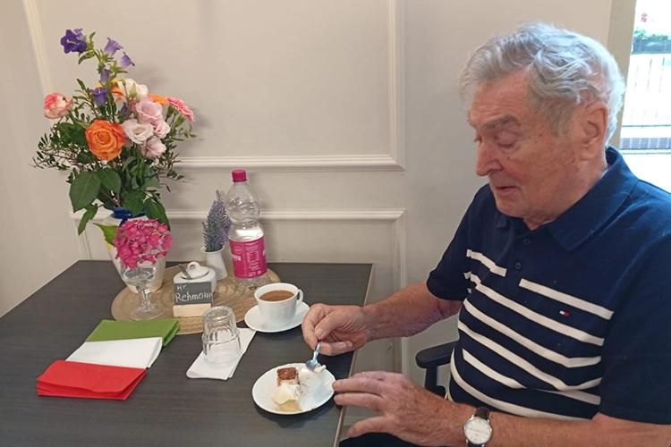 Älterer Mann isst am Tisch Tiramisu, daneben Blumenstrauß und Kaffeetasse