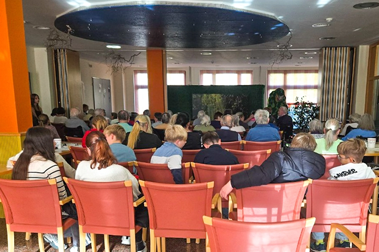 Publikum sitzt in Reihen auf roten Stühlen und schaut auf eine Theaterbühne mit Waldkulisse.