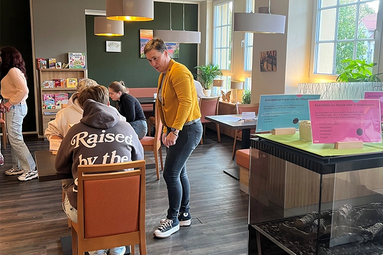 Junge Menschen sitzen ian einem Tisch, eine Frau in gelber Jacke steht erklärend daneben.