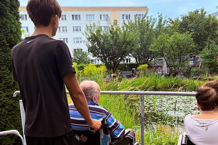 Jugendlicher schiebt älteren Mann im Rollstuhl zu einem Gartenteich.