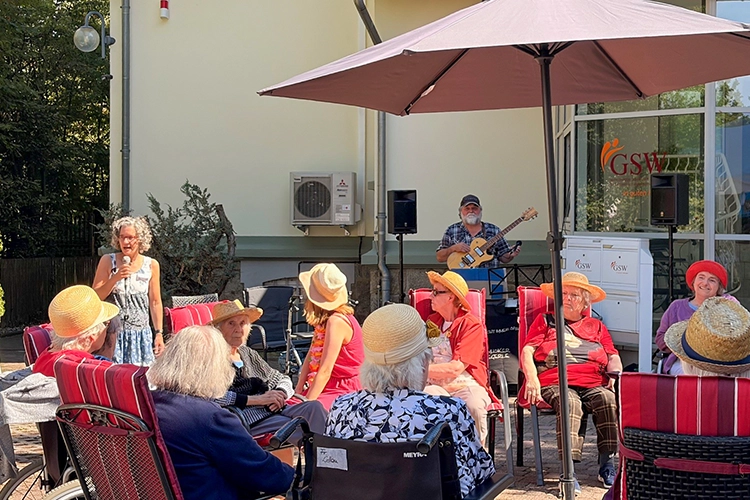 Ältere Menschen mit Strohhüten sitzen auf Terrasse, Sängerin und Gitarrist musizieren.