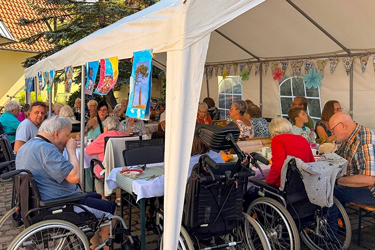 Ältere Menschen in Rollstühlen sitzen unter Festzelt an gedeckten Tischen.