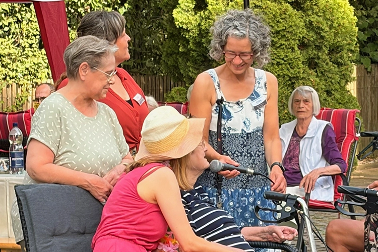 Drei Frauen beugen sich lächelnd zu einer Dame mit Mikrofon auf der Terrasse.