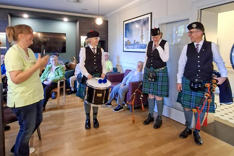 Drei Musiker in Schottenrock mit Dudelsack spielen vor Zuhörern im Aufenthaltsraum.