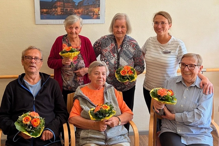 Sechs ältere Personen und eine Pflegerin posieren mit Blumensträußen.
